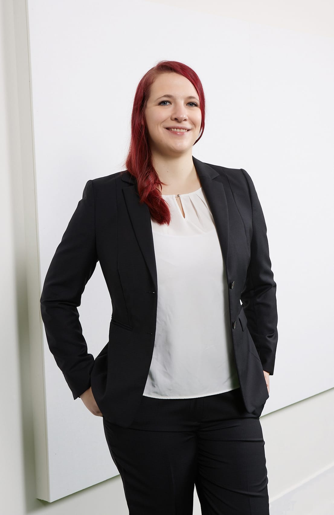 Eine lächelnde Frau mit rotem Haar in einem schwarzen Anzug steht vor einer weißen Wand.