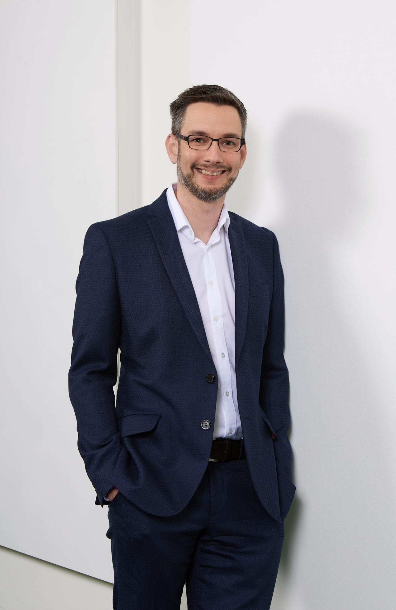 Ein Mann in einem blauen Anzug mit Brille steht lächelnd an einer Wand, die teilweise weiß ist.