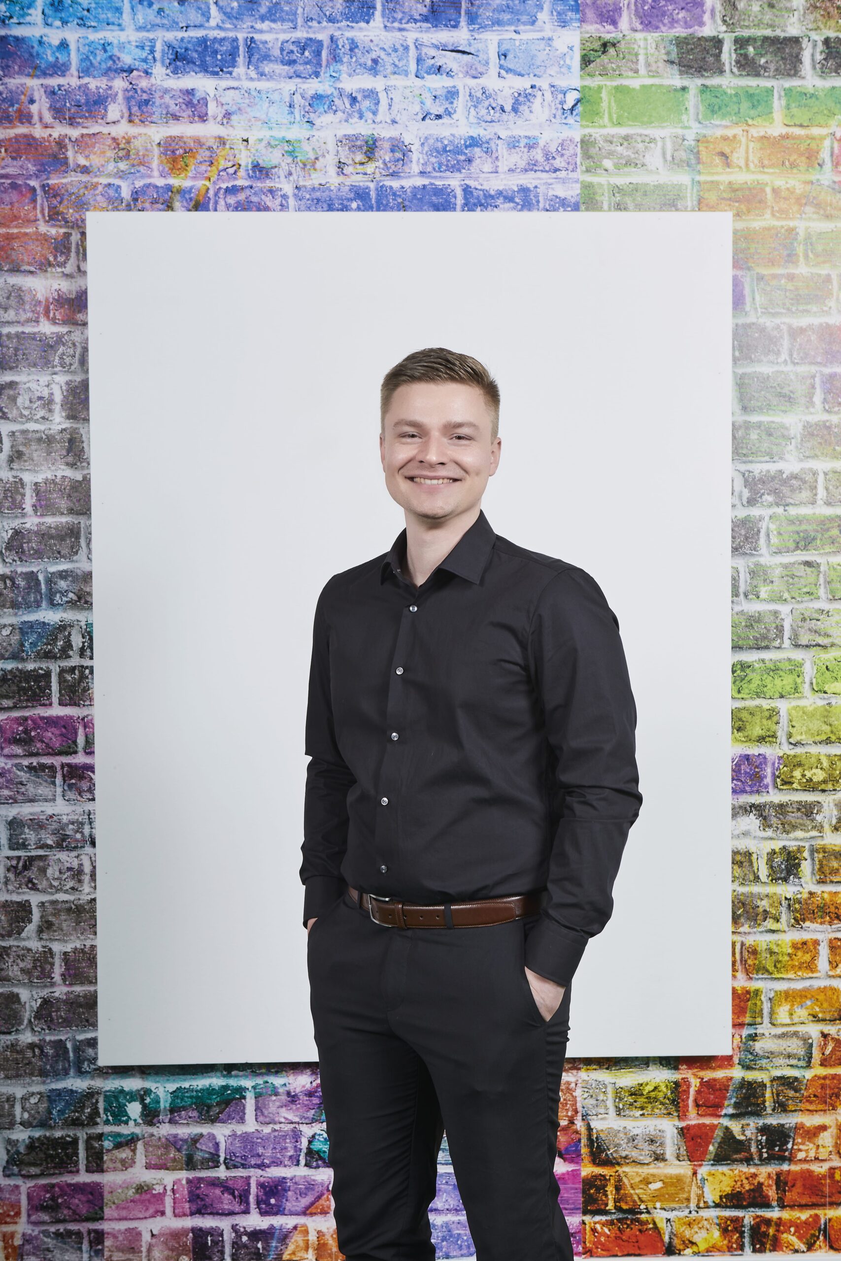 Junger Mann in schwarzem Hemd und Hose steht vor einer wei&szlig;en Leinwand und einer bunten Ziegelwand.
