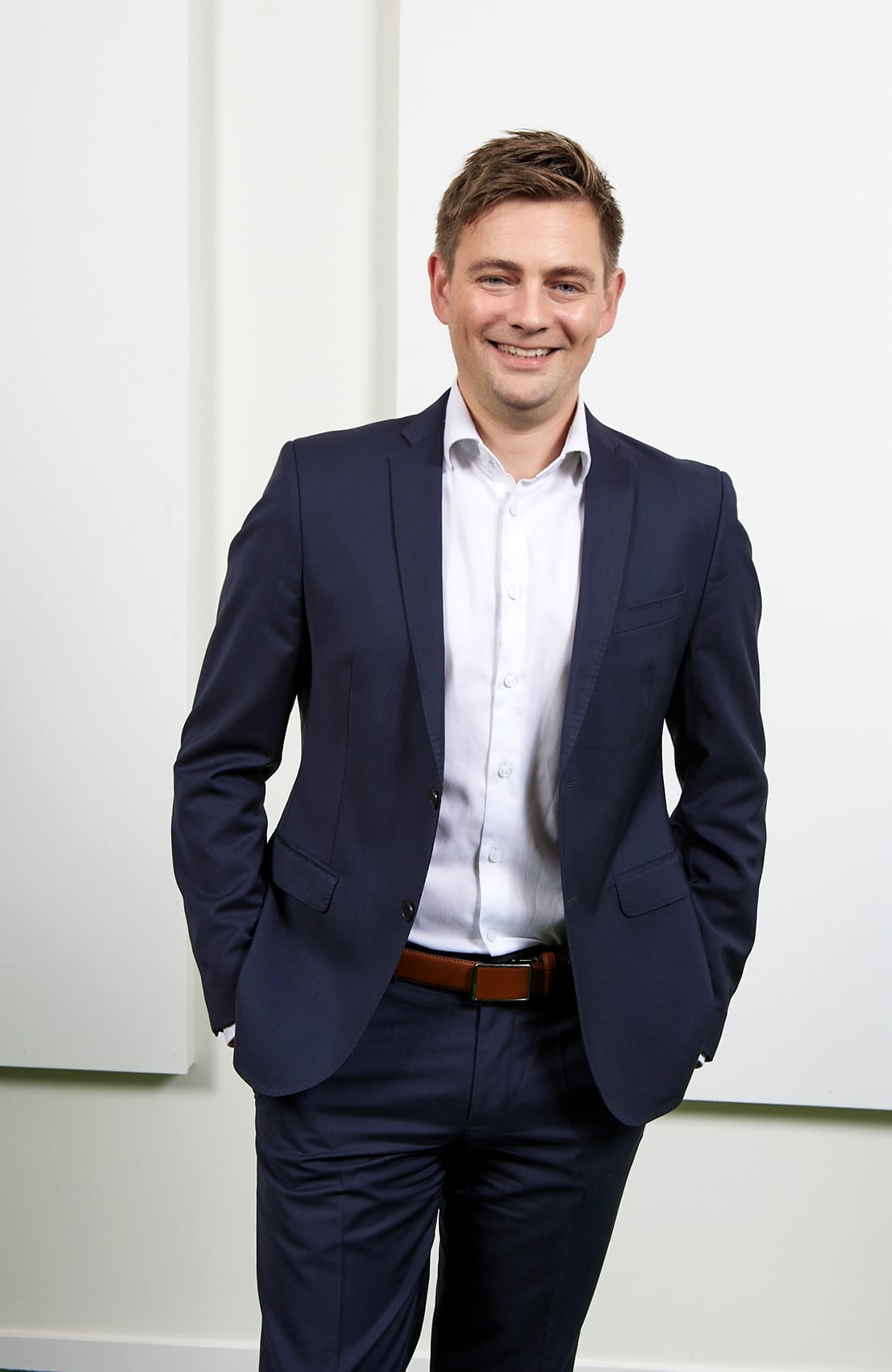 Ein l&auml;chelnder Mann in einem dunkelblauen Anzug steht mit verschr&auml;nkten H&auml;nden vor einer wei&szlig;en Wand.