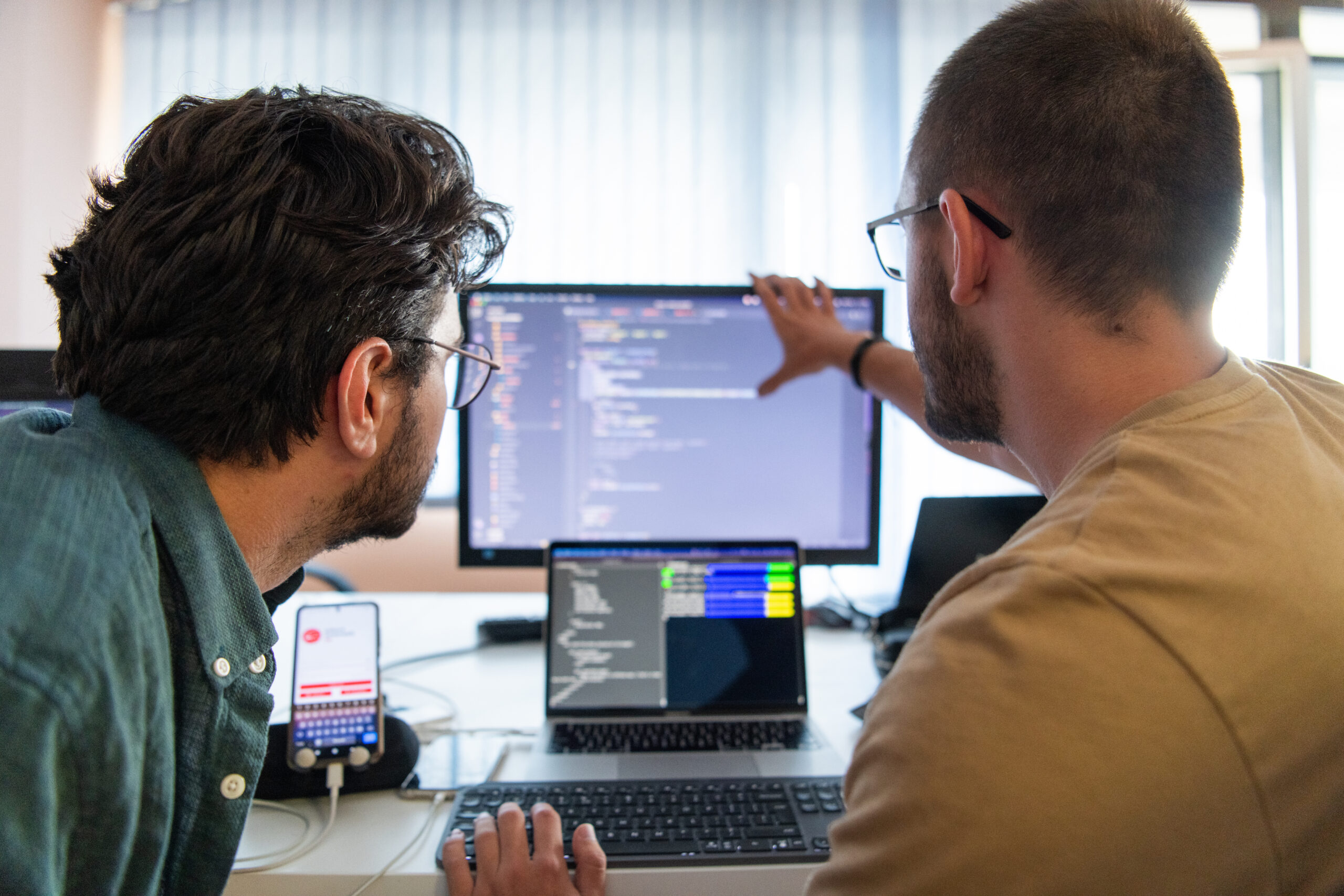 Zwei Personen arbeiten an einem Computer, w&auml;hrend sie Code auf einem Bildschirm besprechen. Ein Smartphone liegt daneben.