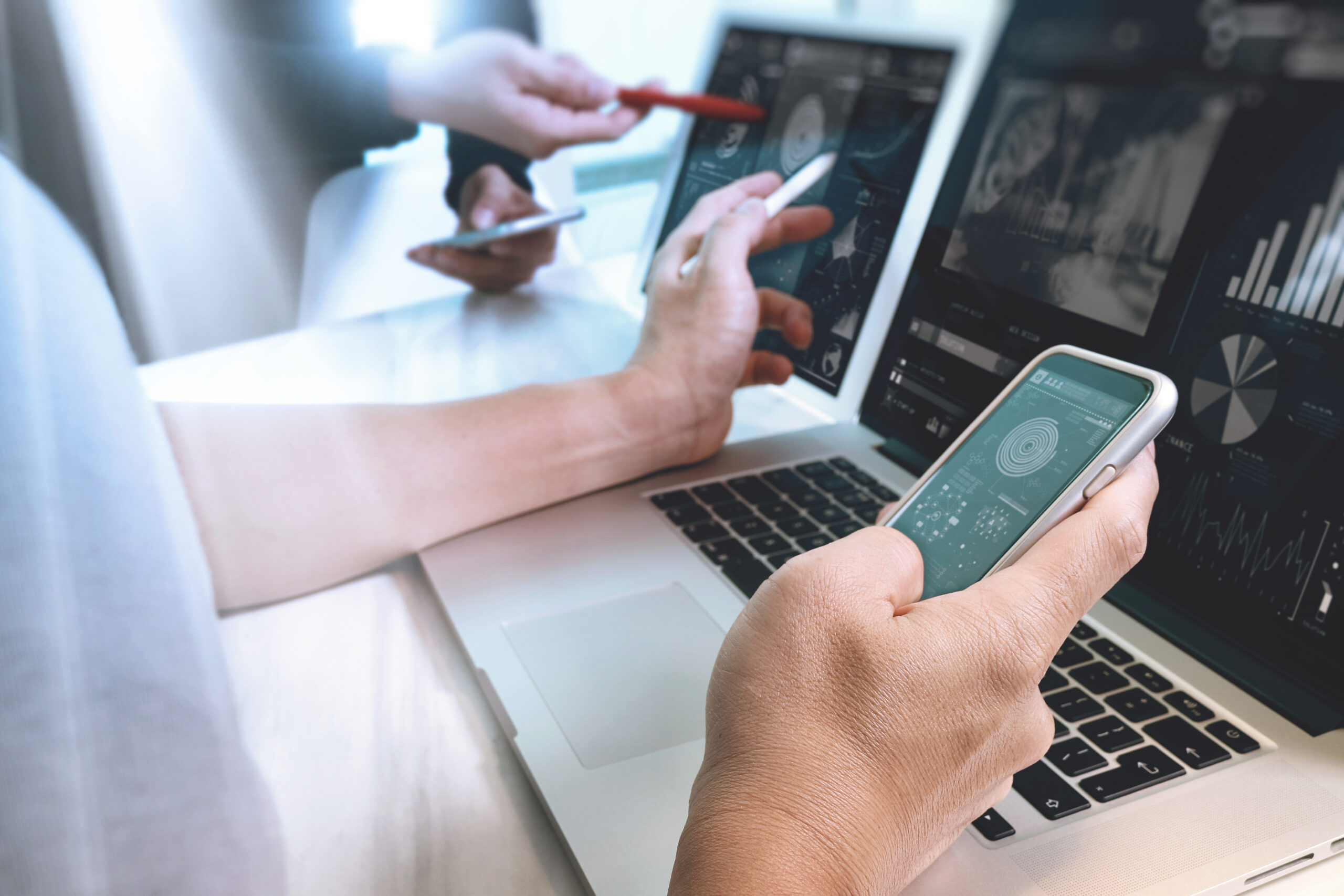 H&auml;nde verwenden Smartphone und Laptop, w&auml;hrend Datenanalysen auf Bildschirmen angezeigt werden.