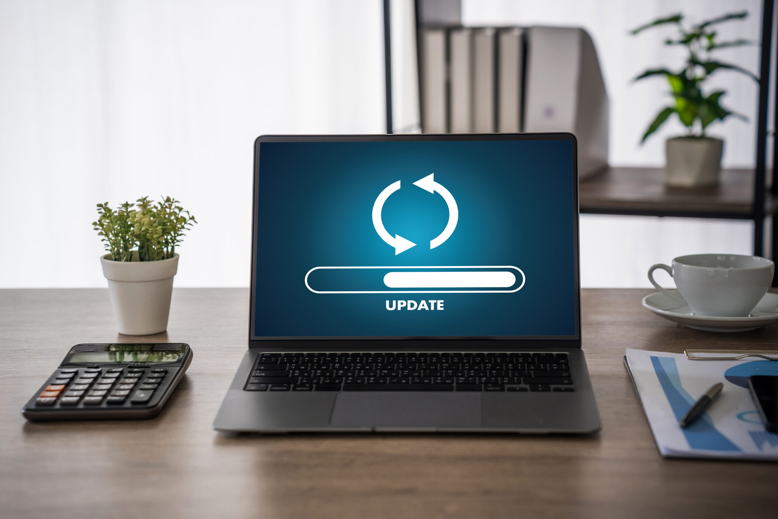 Laptop mit Update-Anzeige auf dem Bildschirm, umgeben von einer Pflanze, einer Tasse und einem Taschenrechner auf einem Tisch.