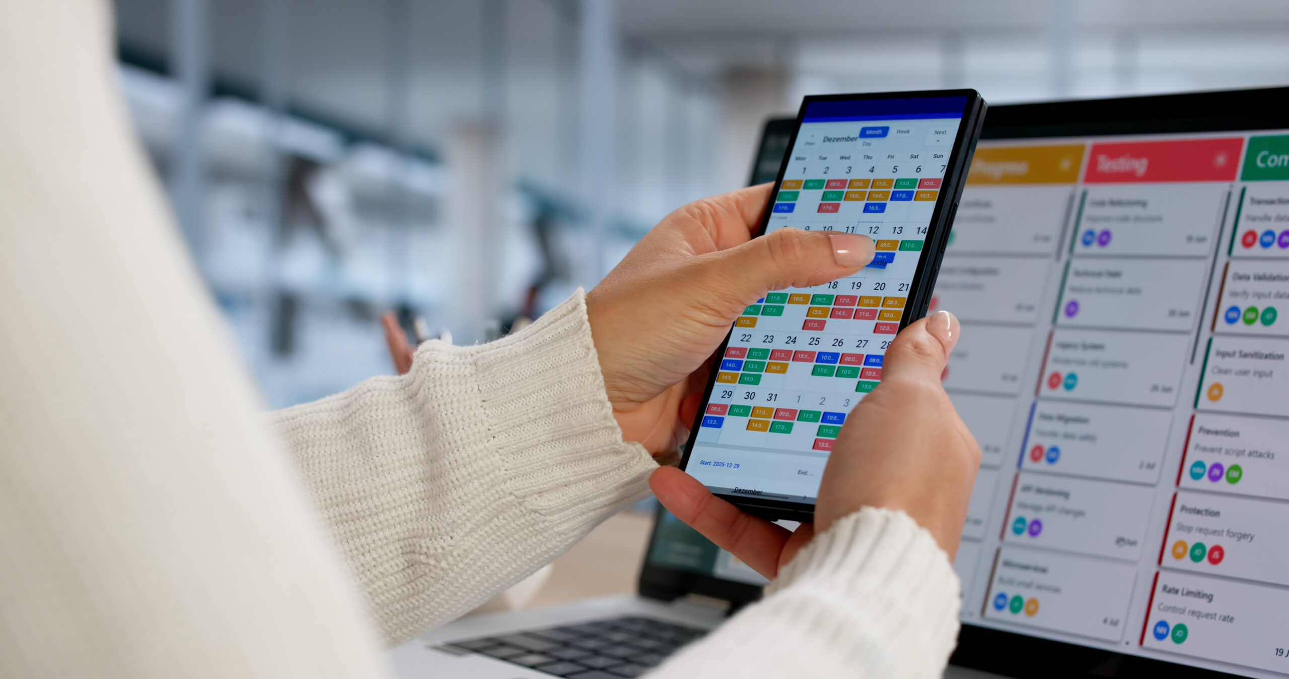 Eine Person &uuml;berpr&uuml;ft einen Kalender auf einem Smartphone, w&auml;hrend ein Computerbildschirm mit Aufgaben und Informationen sichtbar ist.