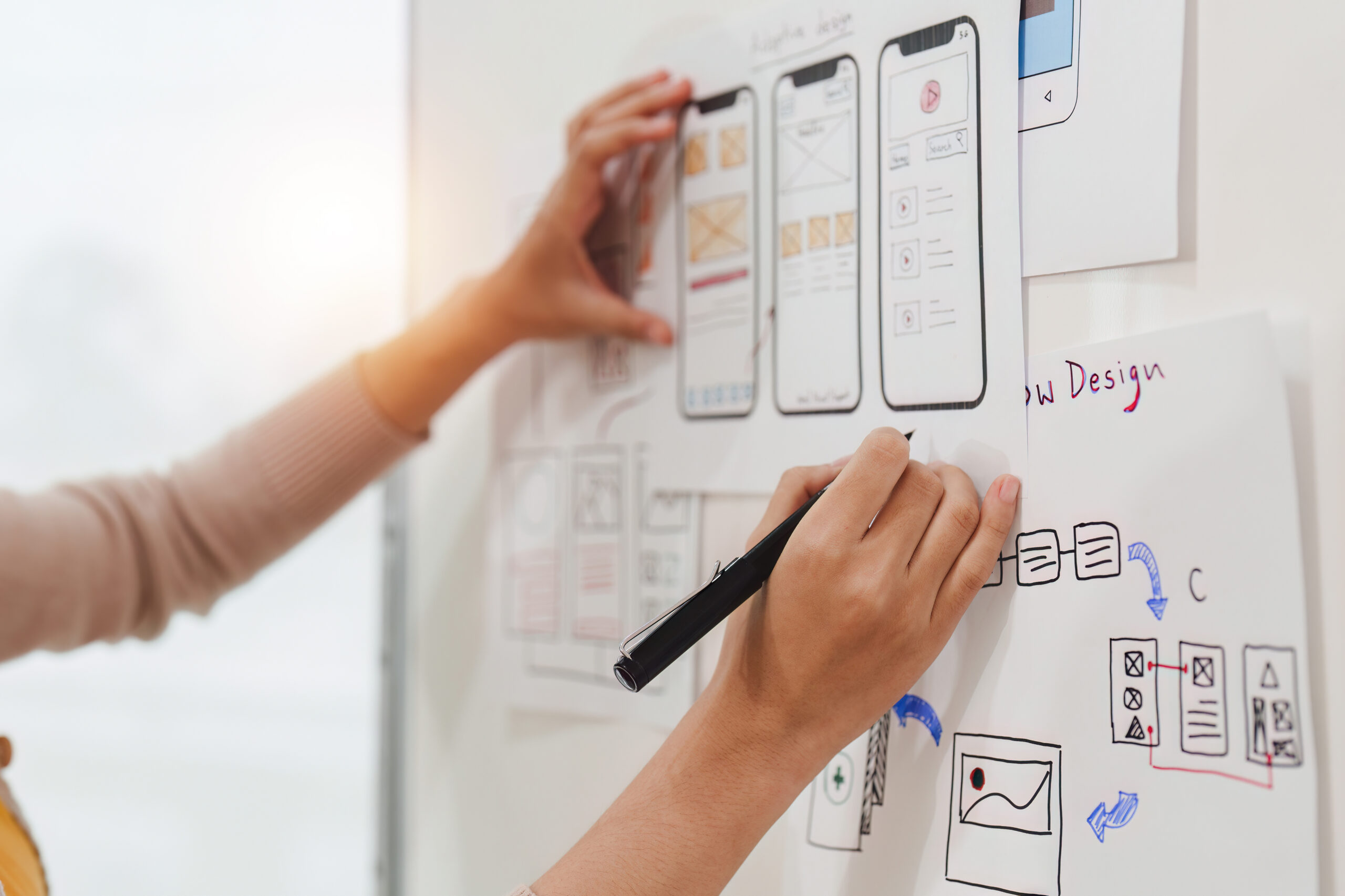 Eine Hand skizziert auf einem Papier, umgeben von Entw&uuml;rfen f&uuml;r mobile App-Layouts an einer Wand.